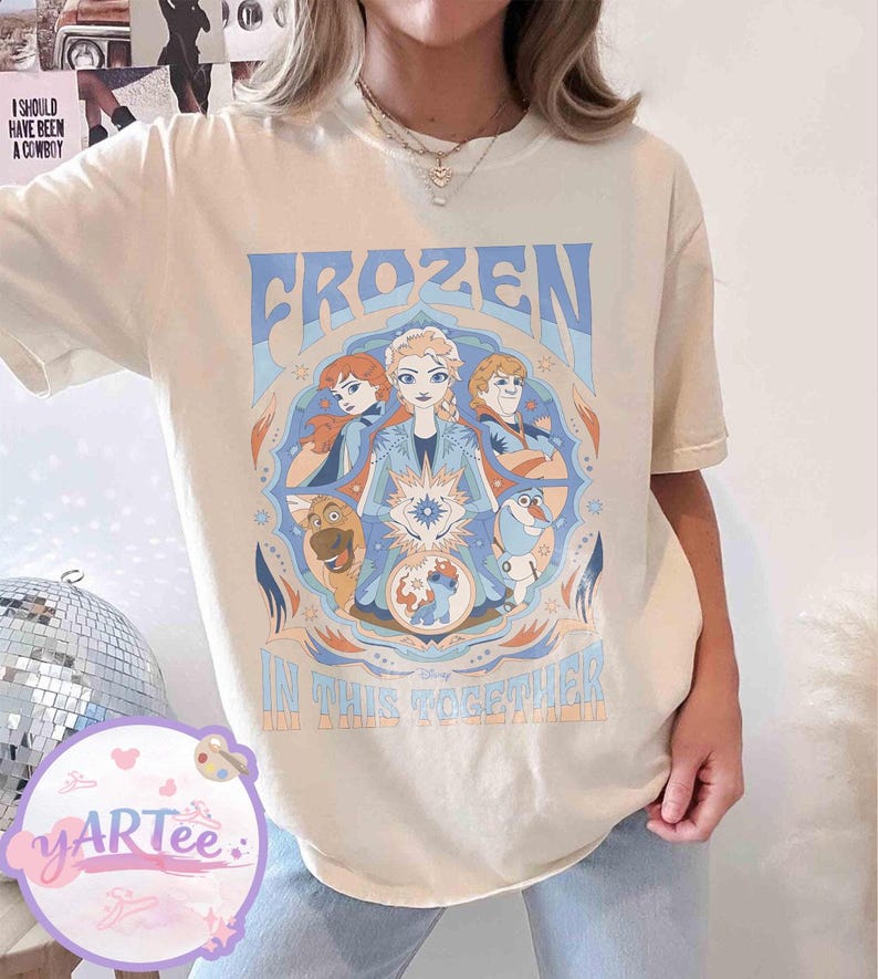 May include: Cream-coloured t-shirt with a Frozen movie graphic. The design features the word "FROZEN" in blue, character illustrations, and the phrase "IN THIS TOGETHER". The shirt has short sleeves.
