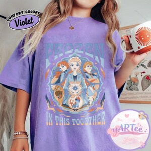 May include: A violet t-shirt with a graphic design from the film "Frozen." The design features the word "FROZEN" in blue, alongside images of the characters Anna, Elsa, Kristoff, and Olaf. The phrase "IN THIS TOGETHER" is also visible.