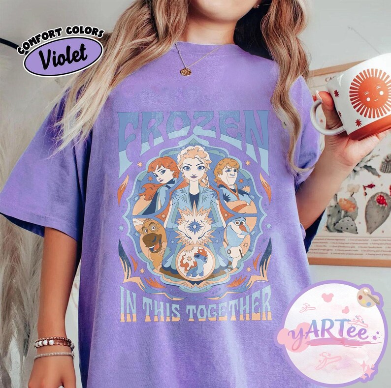 May include: A violet t-shirt with a graphic design from the film "Frozen." The design features the word "FROZEN" in blue, alongside images of the characters Anna, Elsa, Kristoff, and Olaf. The phrase "IN THIS TOGETHER" is also visible.