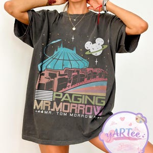 May include: Dark grey t-shirt with a colourful graphic of a futuristic building, a planet with Mickey Mouse ears, and the text "Paging Mr. Morrow". The shirt has a vintage, distressed appearance.