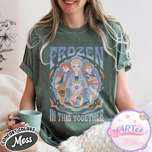 May include: Moss green t-shirt with the word "FROZEN" above a graphic of characters from the film Frozen. The phrase "IN THIS TOGETHER" is below the graphic. The shirt has rolled-up sleeves.