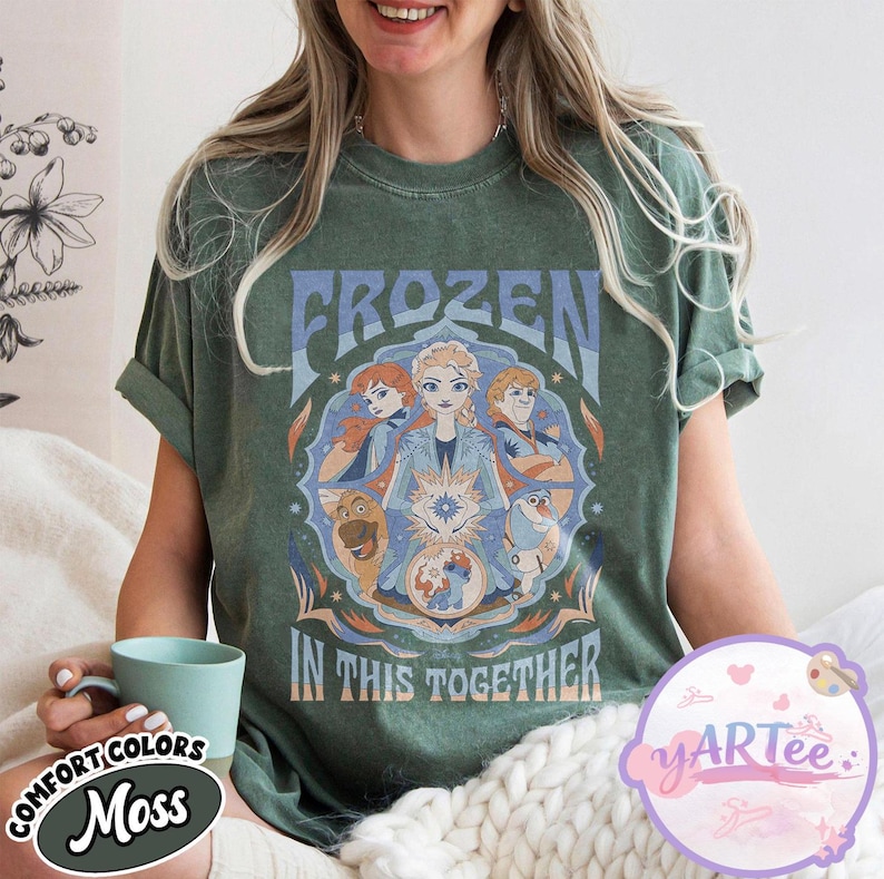 May include: Moss green t-shirt with the word "FROZEN" above a graphic of characters from the film Frozen. The phrase "IN THIS TOGETHER" is below the graphic. The shirt has rolled-up sleeves.