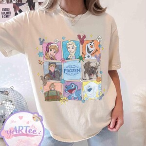 May include: Cream-colored t-shirt featuring a colorful graphic of characters from the Disney movie "Frozen." The design includes images of Anna, Elsa, Olaf, Kristoff, Sven, and other characters, surrounded by floral and decorative elements. The word "FROZEN" is also displayed.