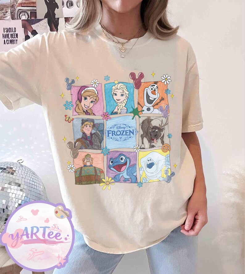 May include: Cream-colored t-shirt featuring a colorful graphic of characters from the Disney movie "Frozen." The design includes images of Anna, Elsa, Olaf, Kristoff, Sven, and other characters, surrounded by floral and decorative elements. The word "FROZEN" is also displayed.