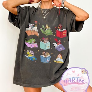 Puede incluir: Camiseta gris oscuro con un dise&ntilde;o caprichoso de personajes animados leyendo libros. El gr&aacute;fico incluye personajes como un gato, un gallo, una rana, un drag&oacute;n y un rat&oacute;n, cada uno absorto en un libro colorido. La camiseta tiene un corte holgado.
