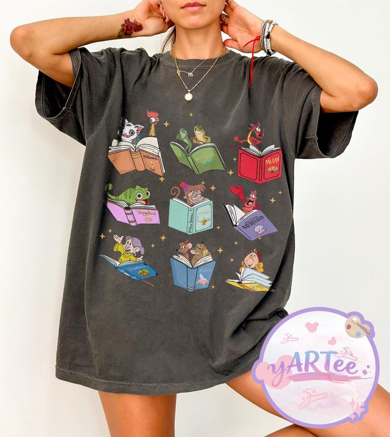 Puede incluir: Camiseta gris oscuro con un dise&ntilde;o caprichoso de personajes animados leyendo libros. El gr&aacute;fico incluye personajes como un gato, un gallo, una rana, un drag&oacute;n y un rat&oacute;n, cada uno absorto en un libro colorido. La camiseta tiene un corte holgado.