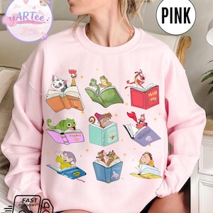 Puede incluir: Sudadera rosa claro con personajes de dibujos animados leyendo libros. Los personajes incluyen una rana, un lagarto y un cangrejo. Los libros tienen t&iacute;tulos como "Moana" y "Mulan". La palabra "PINK" est&aacute; en un c&iacute;rculo blanco en la parte superior derecha.