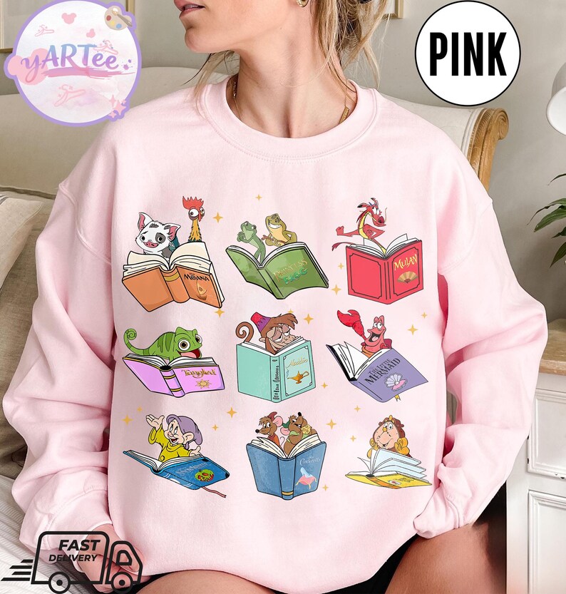 Puede incluir: Sudadera rosa claro con personajes de dibujos animados leyendo libros. Los personajes incluyen una rana, un lagarto y un cangrejo. Los libros tienen t&iacute;tulos como "Moana" y "Mulan". La palabra "PINK" est&aacute; en un c&iacute;rculo blanco en la parte superior derecha.