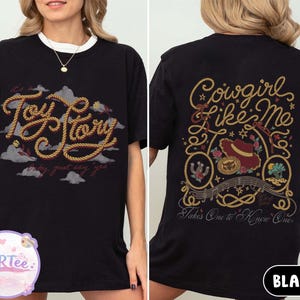 May include: A black t-shirt featuring a front graphic with the text Toy Story in a rope-style font against a background of clouds. The back graphic displays the text Cowgirl Like Me and Takes One to Know One with a cowboy hat and rope motifs.