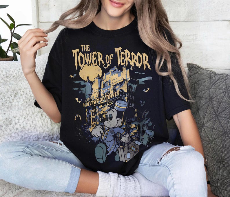 May include: Black t-shirt featuring a graphic of Mickey Mouse in a bellhop uniform in front of the Hollywood Tower Hotel. The text "The Tower of Terror" is printed above the image in a distressed font. The shirt is a relaxed fit.