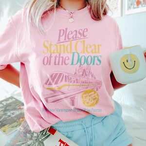 May include: Pink t-shirt with the text "Please Stand Clear of the Doors" in a colorful font, featuring a monorail and castle graphic. A white mug with a yellow smiley face is held in the hand. The shirt is paired with light blue shorts.