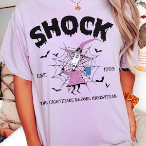 May include: A light purple t-shirt featuring the word "SHOCK" in black, above a graphic of a character from "The Nightmare Before Christmas" inside a spiderweb. The text "EST. 1993" and "THE NIGHTMARE BEFORE CHRISTMAS" are also printed on the shirt.