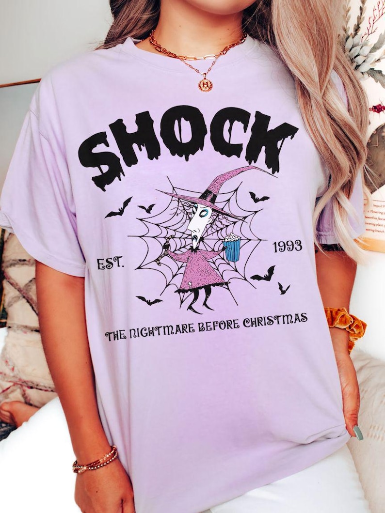 May include: A light purple t-shirt featuring the word "SHOCK" in black, above a graphic of a character from "The Nightmare Before Christmas" inside a spiderweb. The text "EST. 1993" and "THE NIGHTMARE BEFORE CHRISTMAS" are also printed on the shirt.