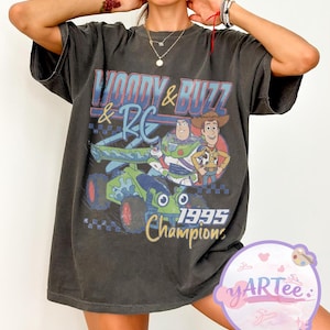 May include: Dark gray t-shirt featuring a graphic of Woody, Buzz Lightyear, and RC from Toy Story. The design includes the text "Woody & Buzz & RC" and "1995 Champions" in blue, red, and yellow. The shirt has short sleeves and a relaxed fit.