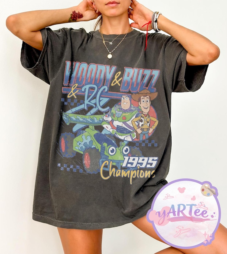 May include: Dark gray t-shirt featuring a graphic of Woody, Buzz Lightyear, and RC from Toy Story. The design includes the text "Woody & Buzz & RC" and "1995 Champions" in blue, red, and yellow. The shirt has short sleeves and a relaxed fit.
