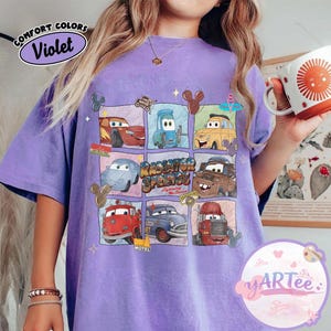 May include: Violet-colored t-shirt featuring a grid of Cars movie characters. The shirt includes the text "Radiator Springs" and "Lightning McQueen." The top left corner has the text "Comfort Colors" and "Violet."