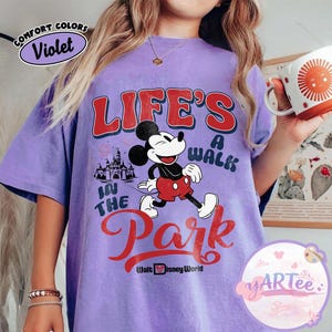 May include: A lilac t-shirt with a Mickey Mouse graphic and the text "LIFE'S A WALK IN THE PARK" in red and white. The design includes a castle and fireworks, with "Walt Disney World" at the bottom. The shirt has a loose fit.