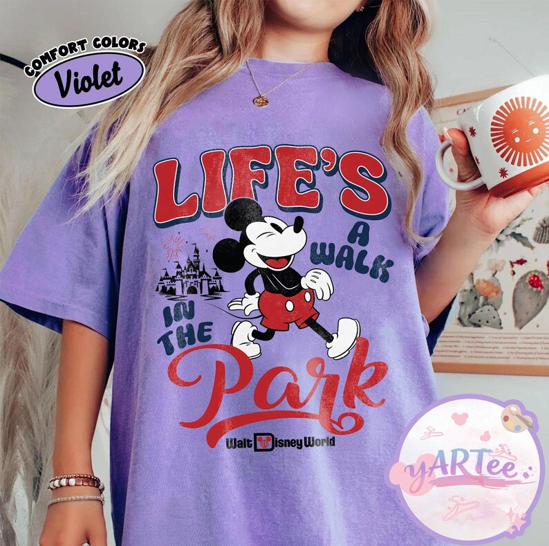 May include: A lilac t-shirt with a Mickey Mouse graphic and the text "LIFE'S A WALK IN THE PARK" in red and white. The design includes a castle and fireworks, with "Walt Disney World" at the bottom. The shirt has a loose fit.