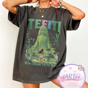 K&ouml;nnte beinhalten: Dunkelgraues T-Shirt mit einer Grafik von Te Fiti aus Moana, mit dem Text "TE FITI" in Gr&uuml;n und Blau. Das Design beinhaltet eine gr&uuml;ne Figur und den Text "The Head Of Te Fiti". Das T-Shirt hat kurze &Auml;rmel.