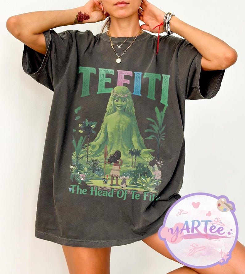K&ouml;nnte beinhalten: Dunkelgraues T-Shirt mit einer Grafik von Te Fiti aus Moana, mit dem Text "TE FITI" in Gr&uuml;n und Blau. Das Design beinhaltet eine gr&uuml;ne Figur und den Text "The Head Of Te Fiti". Das T-Shirt hat kurze &Auml;rmel.