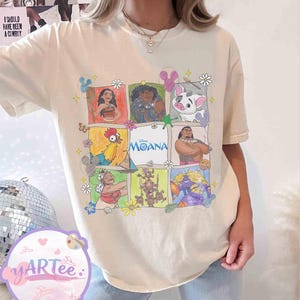 May include: Cream-colored t-shirt featuring a grid of colorful illustrations of characters from the Disney movie "Moana." The word "Moana" is printed in blue. The shirt has short sleeves and a relaxed fit.