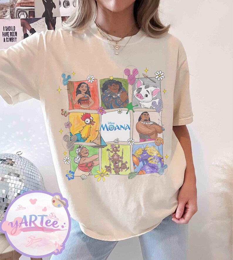 May include: Cream-colored t-shirt featuring a grid of colorful illustrations of characters from the Disney movie "Moana." The word "Moana" is printed in blue. The shirt has short sleeves and a relaxed fit.