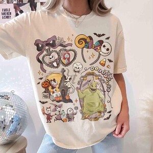 May include: Cream-colored t-shirt featuring a colorful graphic design with characters and elements from "The Nightmare Before Christmas." The design includes Jack Skellington, Sally, Oogie Boogie, and other iconic imagery. The shirt has short sleeves and a crew neck.