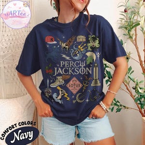 Pu&ograve; includere: T-shirt blu navy con un design grafico con la scritta "PERCY JACKSON" e varie illustrazioni. Il design include un berretto da baseball, Pegaso, un tridente e altri simboli. La maglietta &egrave; etichettata "COMFORT COLORS Navy".
