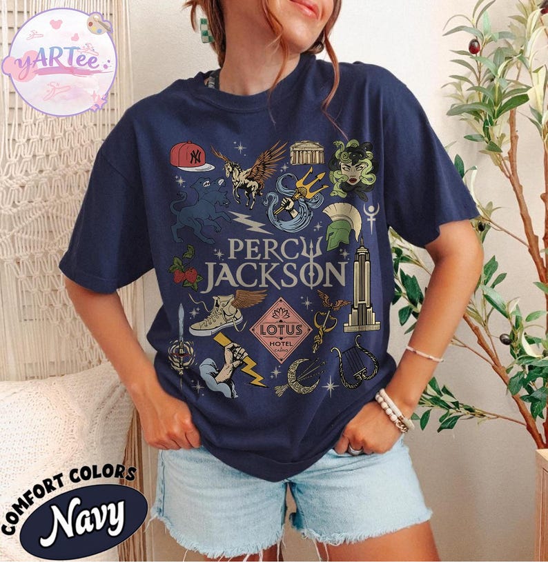 Pu&ograve; includere: T-shirt blu navy con un design grafico con la scritta "PERCY JACKSON" e varie illustrazioni. Il design include un berretto da baseball, Pegaso, un tridente e altri simboli. La maglietta &egrave; etichettata "COMFORT COLORS Navy".