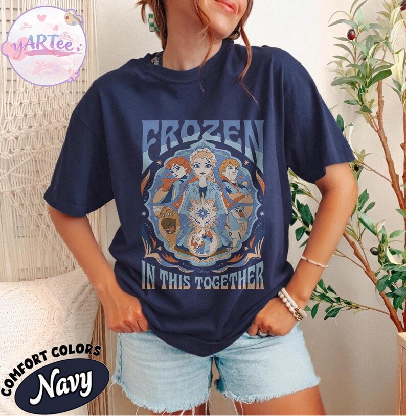 May include: Navy blue t-shirt featuring the text "FROZEN IN THIS TOGETHER" and a graphic of characters from the film Frozen. The shirt has a round neck and short sleeves. The shirt is made of Comfort Colors material.