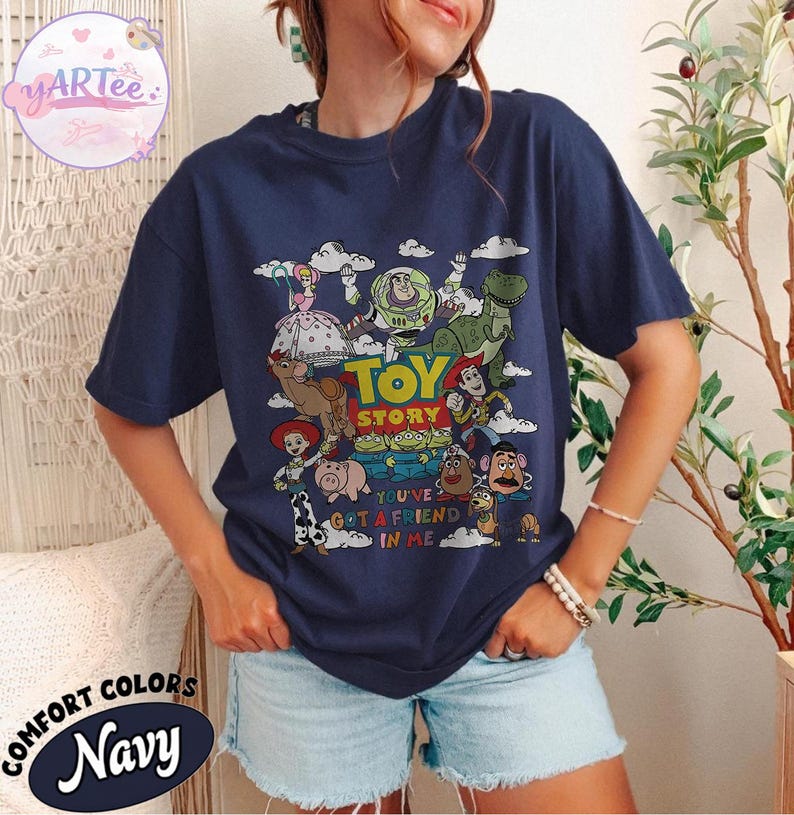 May include: Navy blue t-shirt featuring a colorful graphic of Toy Story characters, including Woody, Buzz Lightyear, and Rex. The text "TOY STORY" and "YOU'VE GOT A FRIEND IN ME" are also displayed. The shirt is labeled "COMFORT COLORS Navy".
