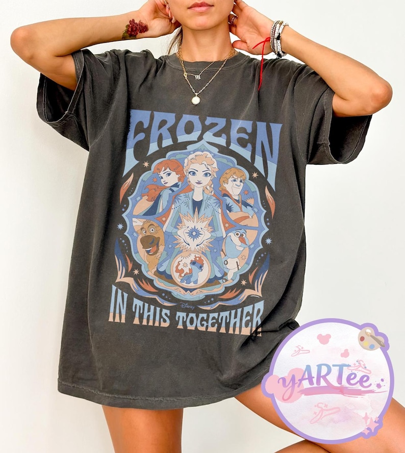 May include: Dark grey t-shirt with a "Frozen" movie graphic. The design includes the text "FROZEN" and "IN THIS TOGETHER" with illustrations of characters from the film. The shirt has short sleeves and a loose fit.