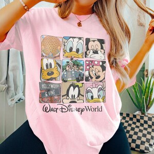 Op de afbeelding: Roze T-shirt met een grafische print van verschillende Disney-figuren, waaronder Mickey Mouse, Donald Duck, Goofy en Pluto. Het shirt heeft ook de tekst "Walt Disney World" en "Hollywood & Vine".