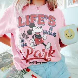 May include: Pale pink t-shirt featuring Mickey Mouse and Cinderella's castle, with the text "LIFE'S A WALK IN THE PARK". The shirt has red and blue lettering. A white mug with a yellow smiley face is held in the hand.