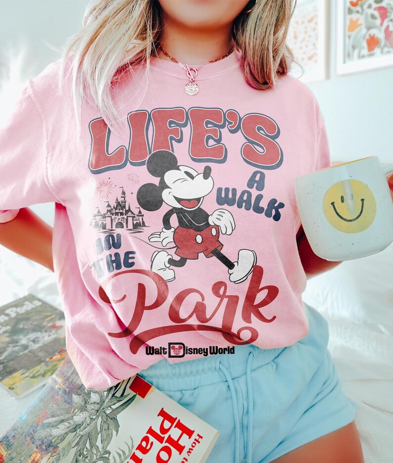 May include: Pale pink t-shirt featuring Mickey Mouse and Cinderella's castle, with the text "LIFE'S A WALK IN THE PARK". The shirt has red and blue lettering. A white mug with a yellow smiley face is held in the hand.