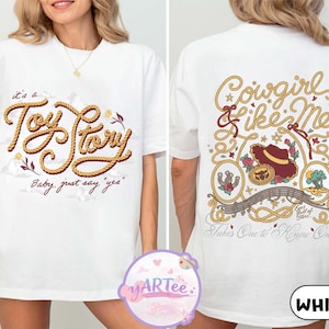 May include: A white t-shirt featuring a front graphic with the text "it's a Toy Story Baby, just say "yes"" in rope-style lettering. The back graphic displays "Cowgirl like Me" and "Takes One to Know One" with illustrations of a hat, horseshoe, and landscape.