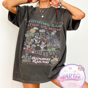 Puede incluir: Camiseta gris oscuro con un gráfico colorido de Mickey y Minnie Mouse, el Pato Donald y otros personajes de dibujos animados en un tren. También se ve el texto "NOTHING CAN STOP US NOW" y "RUNAWAY RAILWAY".