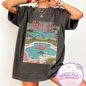 May include: Dark gray t-shirt with a colorful graphic of a pool scene with the text "Bay Lake Florida". The design includes a pool with a Mickey Mouse-shaped float, palm trees, and a castle in the background.