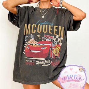 Pu&ograve; includere: T-shirt grigio scuro con Lightning McQueen del film Cars. Il design include il testo "Lightning McQueen" in oro, un'auto da corsa rossa e le frasi "Ultimate Speed" e "Think Fast."