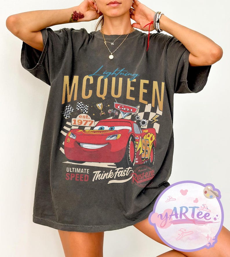 Pu&ograve; includere: T-shirt grigio scuro con Lightning McQueen del film Cars. Il design include il testo "Lightning McQueen" in oro, un'auto da corsa rossa e le frasi "Ultimate Speed" e "Think Fast."