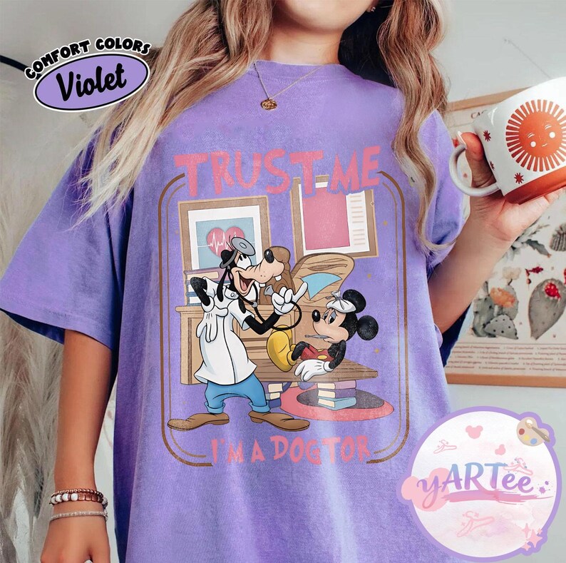 May include: A violet Comfort Colors t-shirt with a cartoon illustration of Goofy and Mickey Mouse in a doctor's office. The text "TRUST ME I'M A DOCTOR" is printed in pink. The shirt has a relaxed fit and is a solid violet colour.