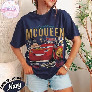 Pu&ograve; includere: T-shirt blu navy con grafica di Saetta McQueen del film Cars. Il design include il testo "Lightning McQueen", "Cars", "est. 1977" e "Ultimate Speed Think Fast". La maglietta &egrave; etichettata "Comfort Colors Navy".