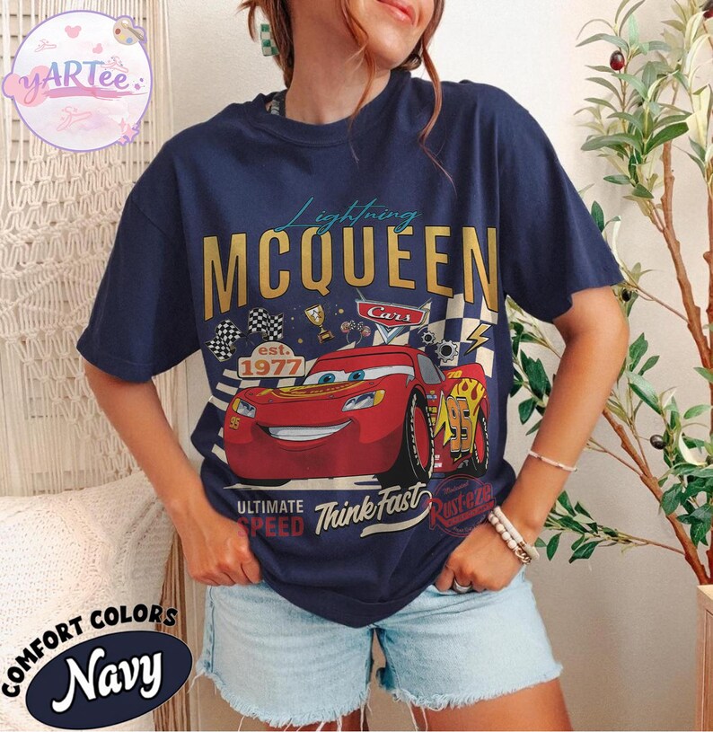 Pu&ograve; includere: T-shirt blu navy con grafica di Saetta McQueen del film Cars. Il design include il testo "Lightning McQueen", "Cars", "est. 1977" e "Ultimate Speed Think Fast". La maglietta &egrave; etichettata "Comfort Colors Navy".
