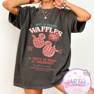 Puede incluir: Camiseta gris oscuro con un dise&ntilde;o gr&aacute;fico retro. El dise&ntilde;o presenta el texto "HOT N' FRESH WAFFLES" con gofres con forma de Mickey Mouse en rojo. El texto adicional incluye "Fresher Than Fresh" y "A Taste of Magic at Breakfast."
