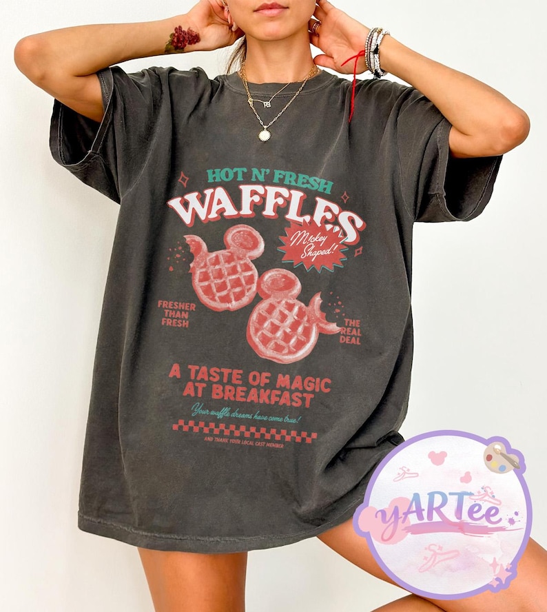 Puede incluir: Camiseta gris oscuro con un dise&ntilde;o gr&aacute;fico retro. El dise&ntilde;o presenta el texto "HOT N' FRESH WAFFLES" con gofres con forma de Mickey Mouse en rojo. El texto adicional incluye "Fresher Than Fresh" y "A Taste of Magic at Breakfast."