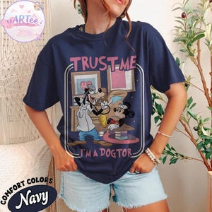 May include: Navy blue t-shirt with a graphic of Goofy as a doctor examining Mickey Mouse. The text "TRUST ME I'M A DOGTOR" is above the graphic. The shirt also has the words "COMFORT COLORS" and "Navy" printed on it.
