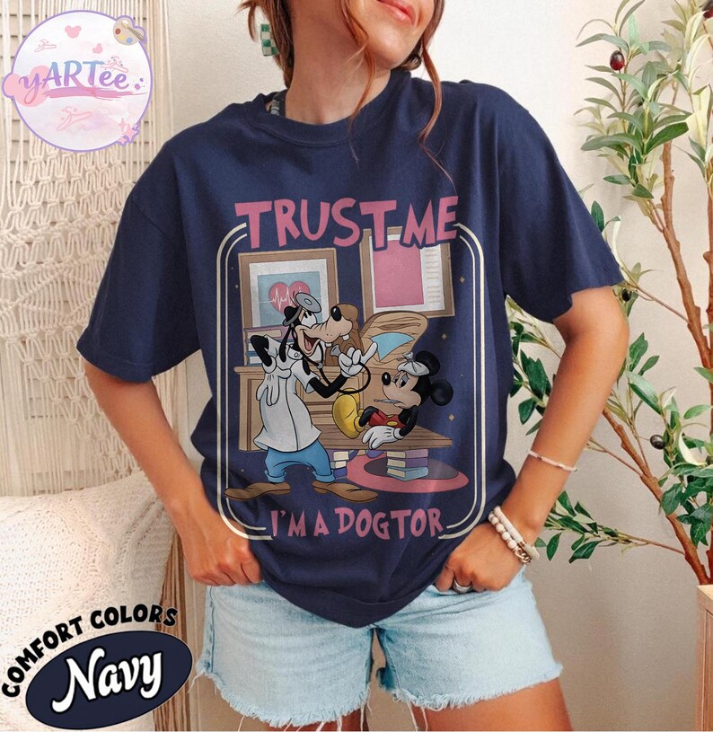 May include: Navy blue t-shirt with a graphic of Goofy as a doctor examining Mickey Mouse. The text "TRUST ME I'M A DOGTOR" is above the graphic. The shirt also has the words "COMFORT COLORS" and "Navy" printed on it.