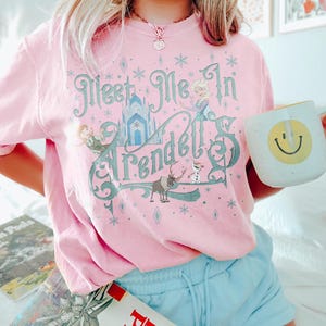 Pode incluir: Camiseta rosa claro com o texto "Meet Me In Arendelle" em verde-azulado e branco, com ilustra&ccedil;&otilde;es de personagens e um castelo. A pessoa est&aacute; segurando uma caneca branca com um rosto sorridente amarelo.