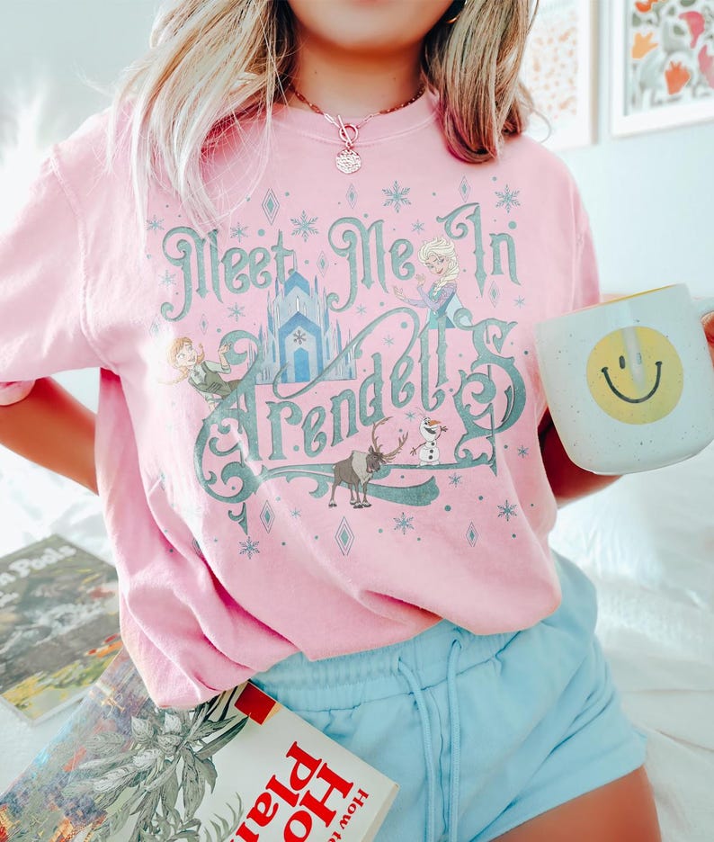 Pode incluir: Camiseta rosa claro com o texto "Meet Me In Arendelle" em verde-azulado e branco, com ilustra&ccedil;&otilde;es de personagens e um castelo. A pessoa est&aacute; segurando uma caneca branca com um rosto sorridente amarelo.