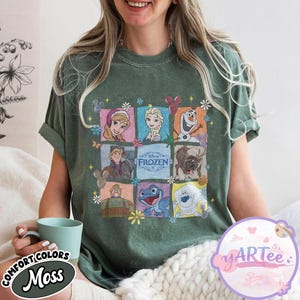 May include: A moss green t-shirt featuring a grid of Disney Frozen characters. The shirt includes images of Anna, Elsa, Olaf, Kristoff, Sven, and other characters. The text "FROZEN" is in the center. The shirt has the text "COMFORT COLORS" and "Moss".